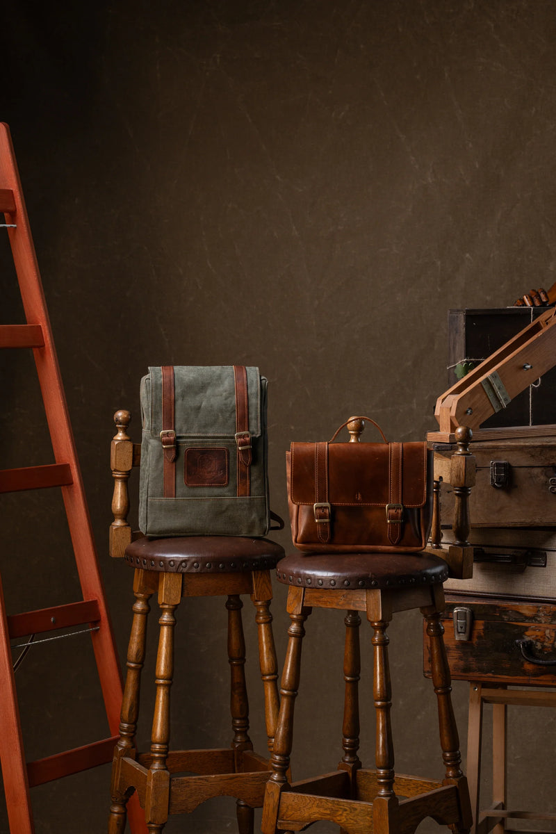 A waxed canvas backpack and a leather briefcase resting on wooden stools