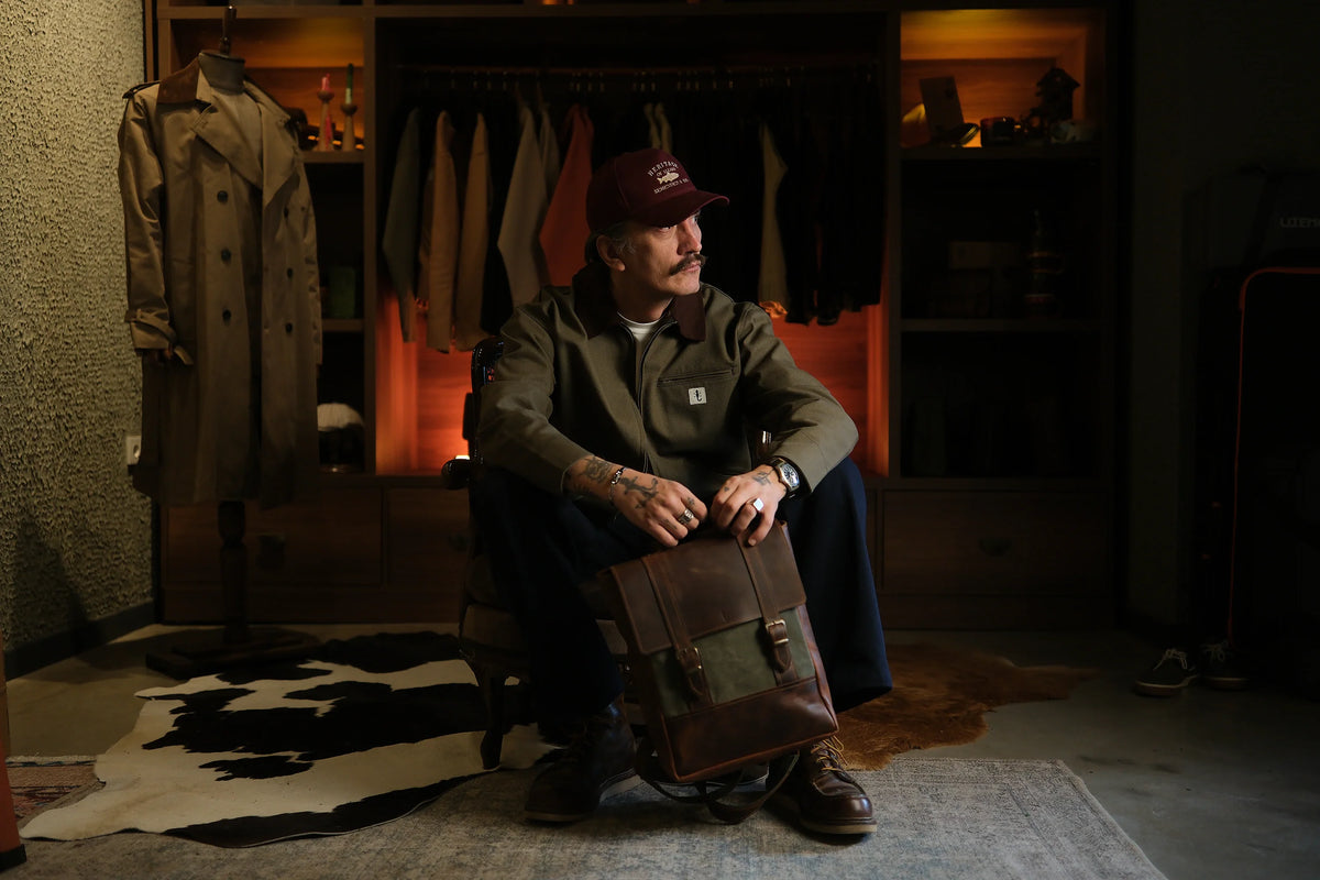 Man in a green work jacket sitting by a wardrobe, a green canvas Honeybag backpack on his lap and a trench coat beside him